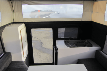 Innenraum eines Campers mit Sitzbank, Tisch, kleinem Kühlschrank und Spüle, durch Fenster Blick auf eine Straße und einen großen Bus im Nebel.