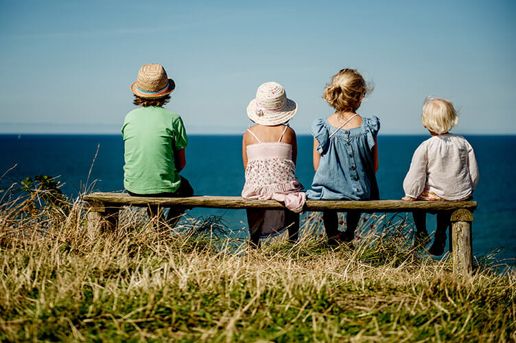 Vier Kinder sitzen auf einer Bank und blicken auf das Meer.
