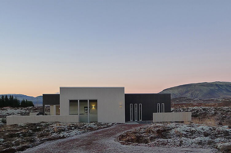 Modernes Ferienhaus mit Flachdach in karger Landschaft im Herbst oder Winter vor Bergen bei Dämmerung.