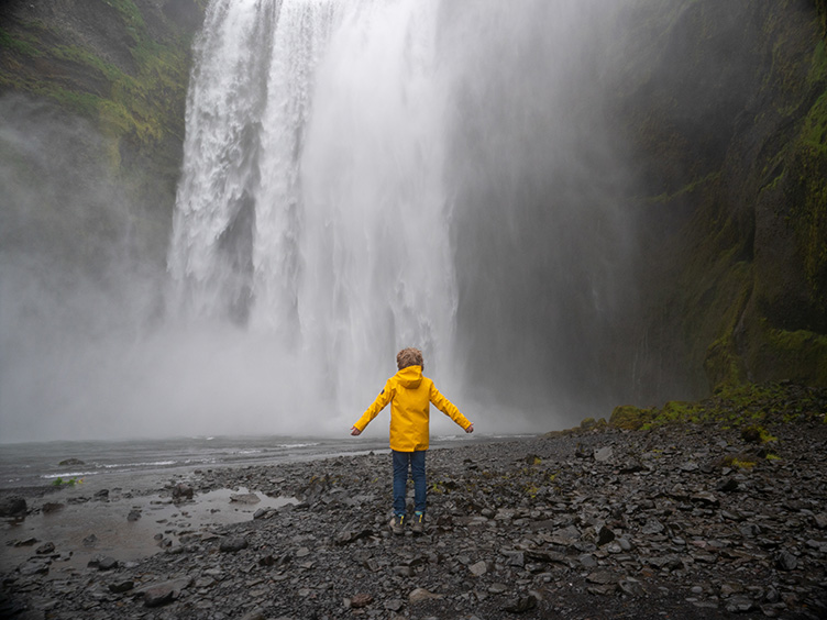 Kind in gelber Regenjacke steht vor einem Wasserfall.
