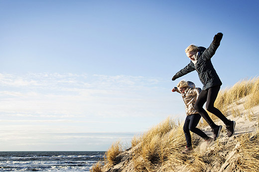 Zwei Kinder springen von einer Düne zum Meer.