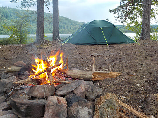 Ein kleines Zelt steht am Wasser hinter einem Lagerfeuer.
