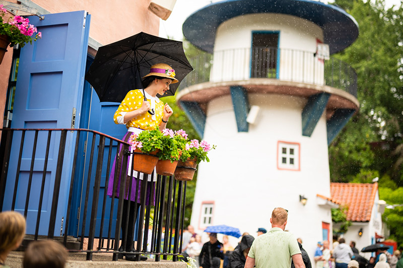 Frau im bunten Kostüm steht auf einem Balkon mit einem schwarzen Regenschirm
