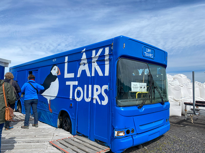 Touristen stehen vor einem blauen Bus mit der Aufschrift Láki Tours und einem aufgemalten Puffin.