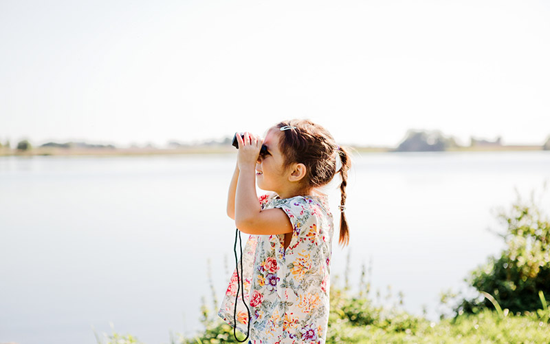 Mädchen steht an einem See und hält ein Fernrohr