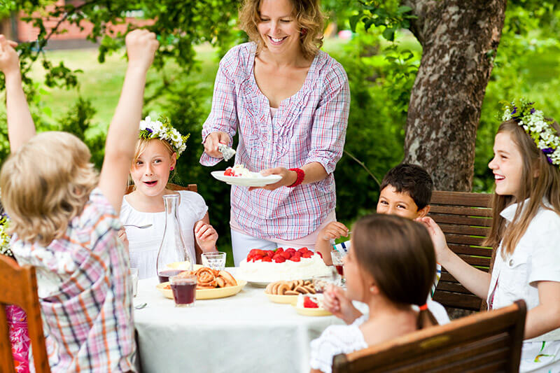 Kinder sitzen um einen Tisch und freuen sich währen eine Frau Torte verteilt.