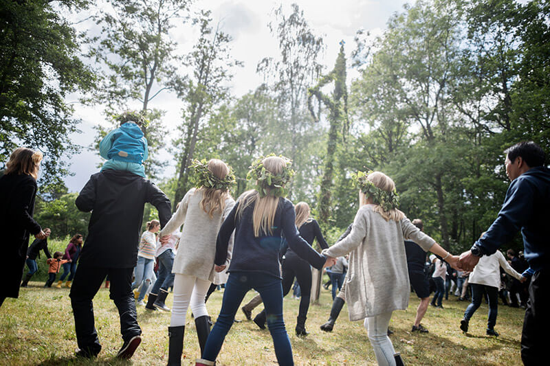 Kinder und Erwachsene tanzen um die Maistange und haben Blumenkränze auf dem Kopf.