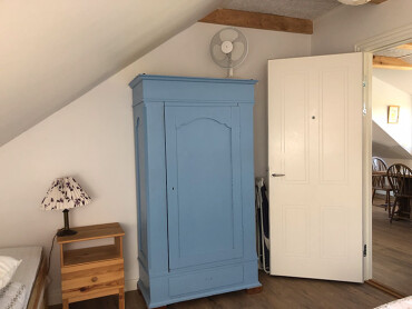 Blick auf den blauen Holzschrank im Schlafzimmer. Durch die offene Tür blickt man ins Esszimmer.