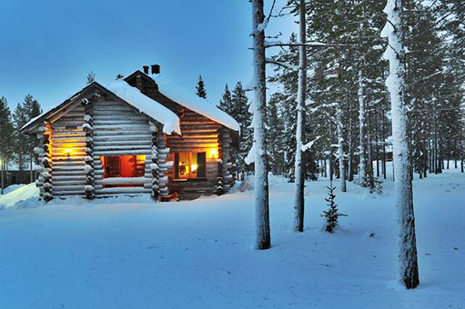 Eine kleine schneebedeckte Hütte steht im Winter am Waldrand und ist beleuchtet.