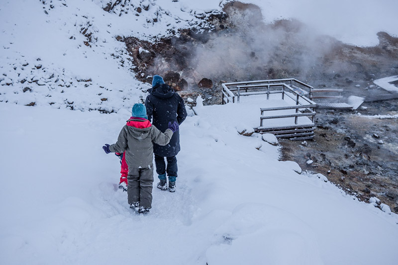 Drei Personen in Winterkleidung laufen über einen verschneiten Pfad zur heißen Quelle.