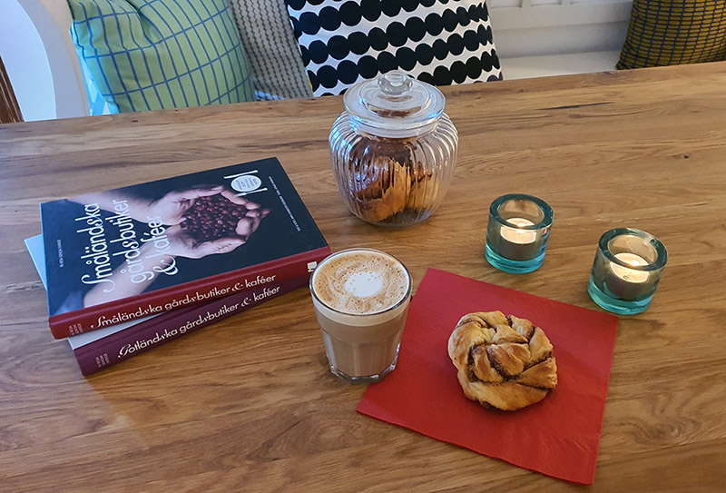 Eine Zimtschnecke und eine Tasse Kaffee stehen auf einem Holztisch. Dahinter steht ein volles Glas mit weiteren Zimtschnecken und zwei Backbücher.