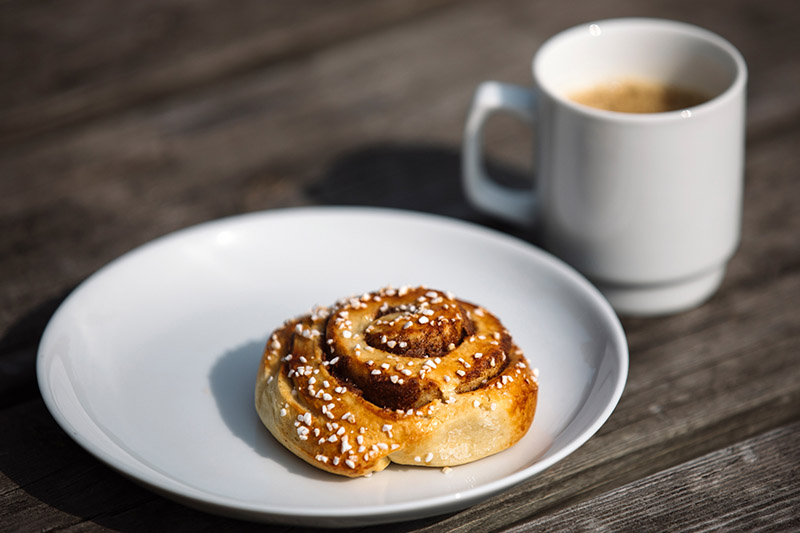 Eine Zimtschnecke ist mit Hagelzucker bestreut. Im Hintergrund steht eine weiße Tasse mit Kaffee.