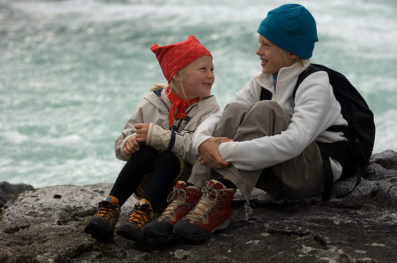 Zwei Kinder in Wanderkleidung sitzen auf einem Felsen am Wasser.