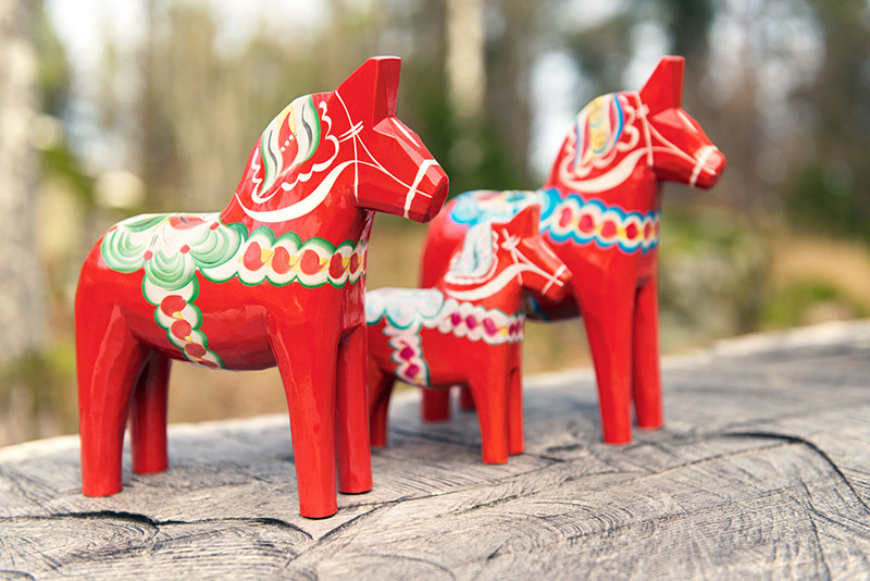 Drei rote Dalapferde stehen auf einem Holzbalken.