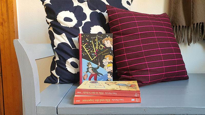 Drei Kinderbücher liegen auf einer Bank. Ein Buch ist angelehnt an bunte Kissen.