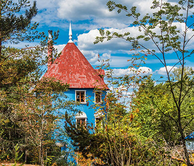 Ein blaues rundes Haus mit rotem Dacht steht versteckt hinter Bäumen.
