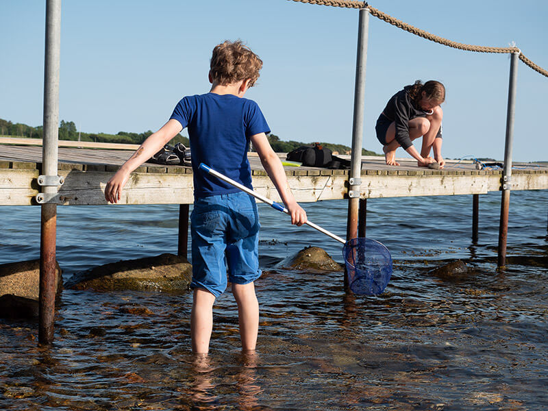Ein Junge steht mit einem Köcher im Wasser vor einem Steg. Auf dem Steg ist ein Mädchen.