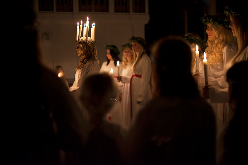 Eine blonde Frau in weißem Gewand und mit Kerzenkranz auf dem Kopf steht vor einer Gruppe von Frauen in weißen Gewändern mit Kerzen.