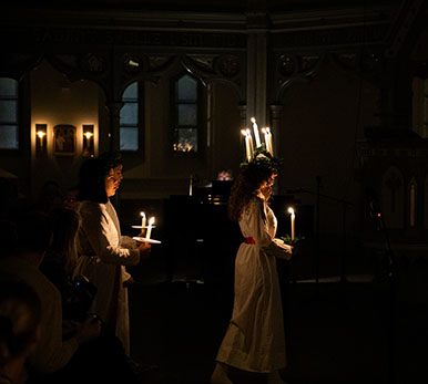 Drei Menschen laufen durch eine dunkle Kirche. Die erste Person hat einen Kranz aus Kerzen auf dem Kopf.