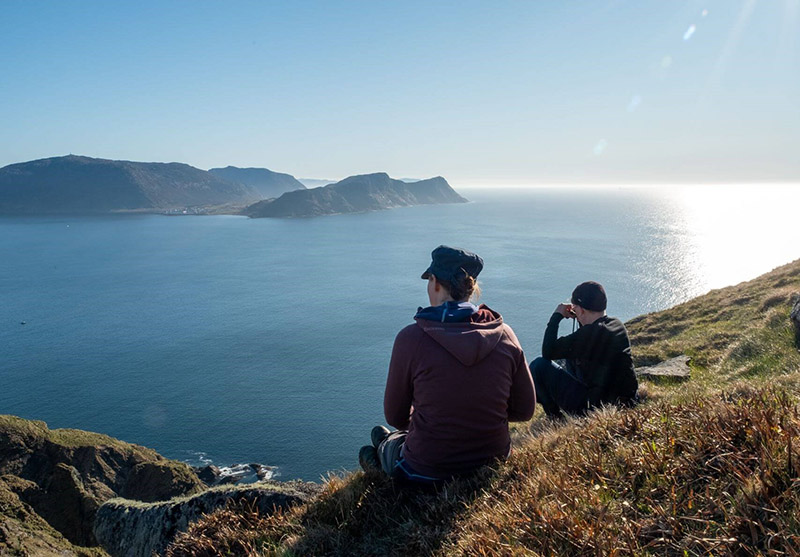 Zwei Wanderer sitzen auf einem grasigen Hang und schauen mit einem Fernglas aufs Meer.