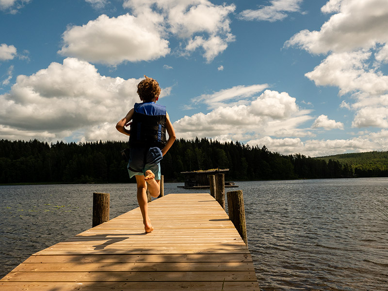 Ein blonder Junge mit Schwimmweste rennt über einen Steg zum Wasser