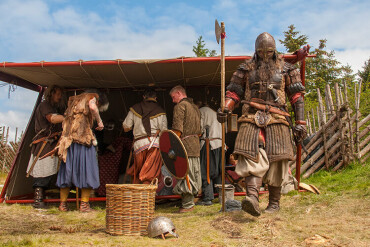 Mehrere Personen in Wikingerkleidung vor einem einfachen Unterstand mit einem Korb und einem Helm auf dem Boden.