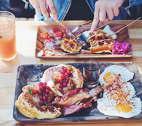 Zwei üppig belegte Schalen mit Waffeln, Eiern und Bacon. Auf der hinteren Schale wird eine Waffel gerade geschnitten. 