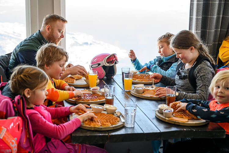 Eine Familie in Winterkleidung sitzt am Tisch und isst ihre Waffeln. Im Hintergrund sieht man einen schneebedeckten Gipfel durchs Fenster. 