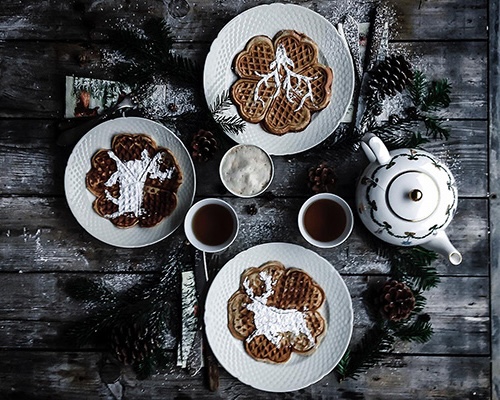 Ein dunkler Holztisch mit weißem Gedeck, auf dem Waffeln mit Puderzucker in Form von Rentieren bestreut sind. 