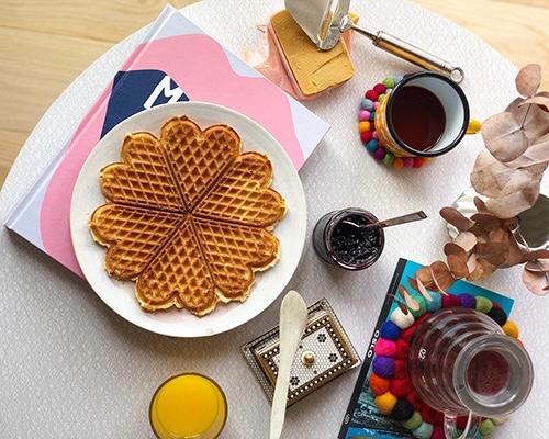 Ein gedeckter Tisch mit Tassen, bunten Untersetzern, einem Teller mit einer Waffel und Braunkäse samt Käsehobel.