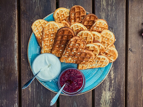 Ein blauer Teller auf dem herzförmige Waffeln liegen. Daneben eine Schale mit Marmelade und saurer Sahne.