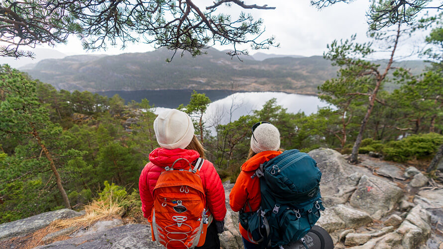 Zwei Frauen in Wanderkleidung mit Wanderrucksäcken stehen auf einem Felsvorsprung und schauen ins Tal. Unten ist ein See und Hügel zu sehen. 