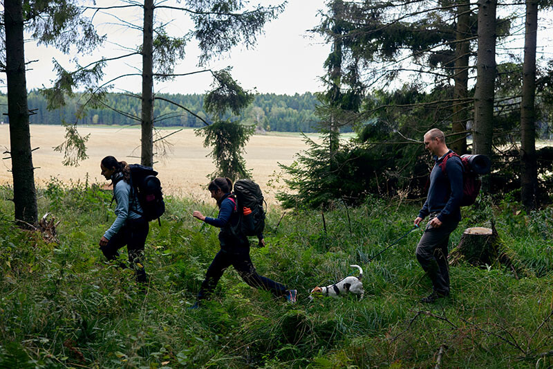 Drei Wanderer mit einem kleinen Hund laufen durch den Wald. Im Hintergrund sieht man einen See.