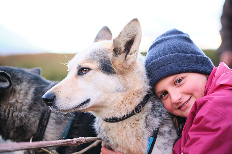 Ein Mädchen mit dunkler Mütze und rosa Jacke umarmt einen blond-grauen Hund.