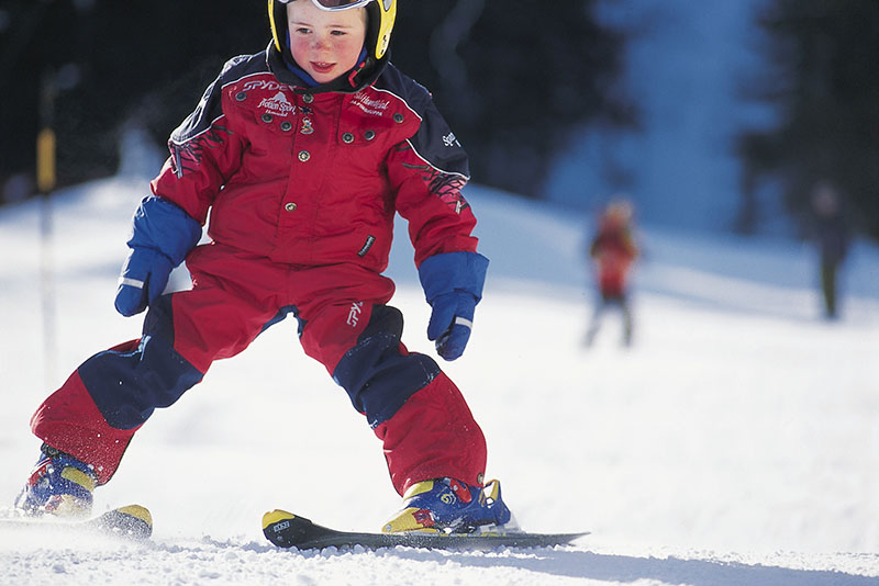 Kleines Kind im roten Schneeanzug steht auf einer Skipiste.