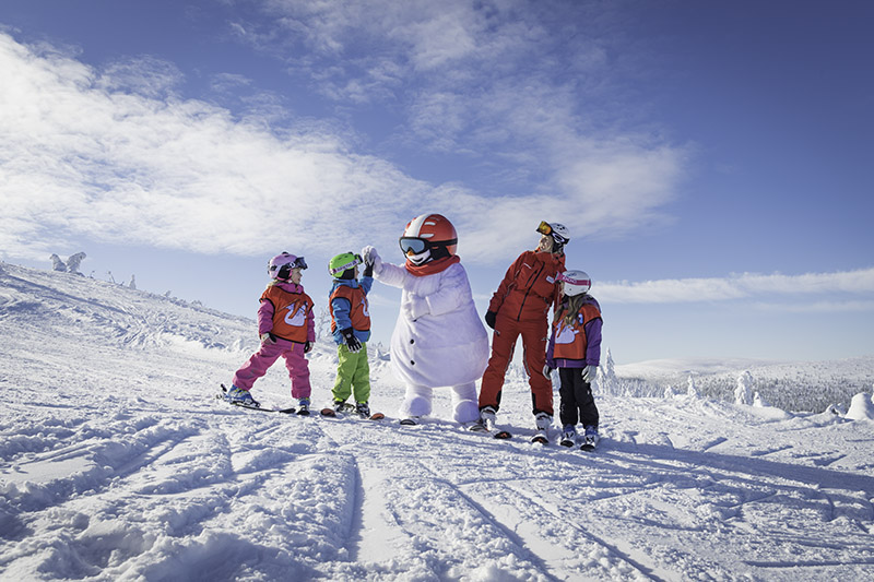 Eine Gruppe Kinder auf Skiern steht auf der Piste mit dem Schneemann Valle. Ein Kind gibt Valle ein High-Five.