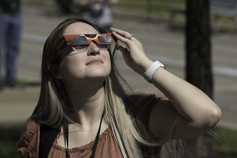 Eine junge Frau hält sich eine rote Papiersonnenbrille für die Sonnenfinsternis vor das Gesicht und schaut in den Himmel.