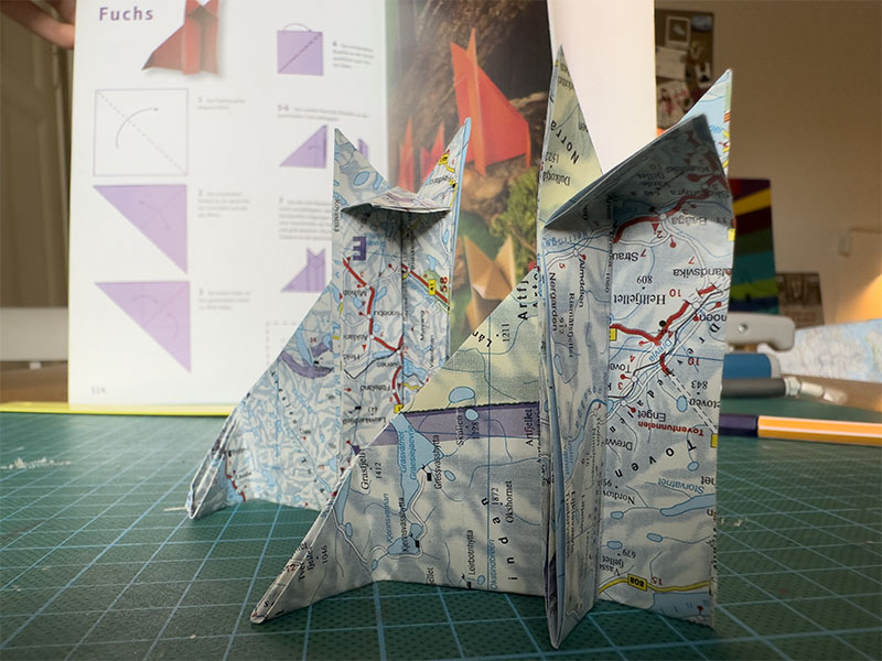 Zwei Origamifüchse aus alten Landkarten. Im Hintergrund ist die Bastelanleitung zu sehen.
