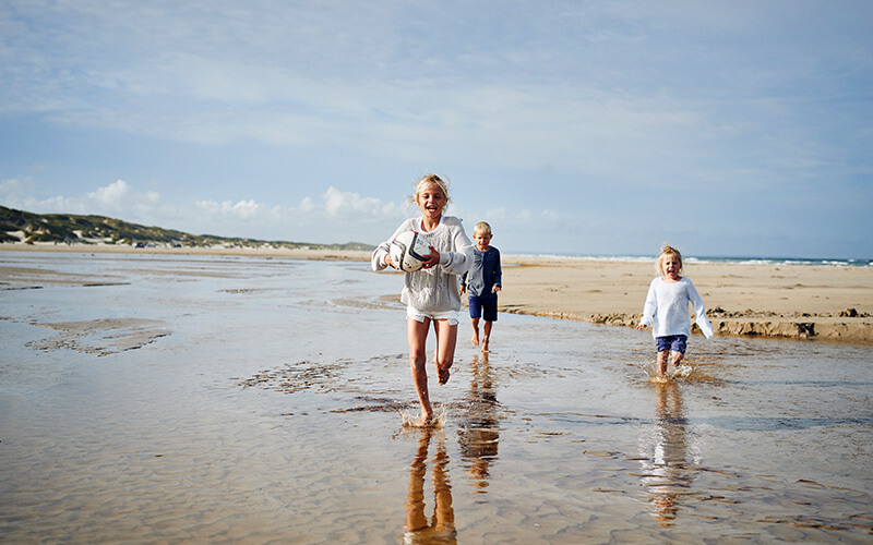Drei Kinder laufen barfuß am Strand, eines hält einen Ball in den Händen.