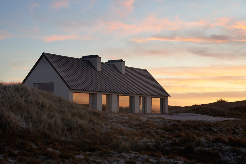 Modernes Haus mit großen Fenstern zwischen Dünen bei Sonnenuntergang.