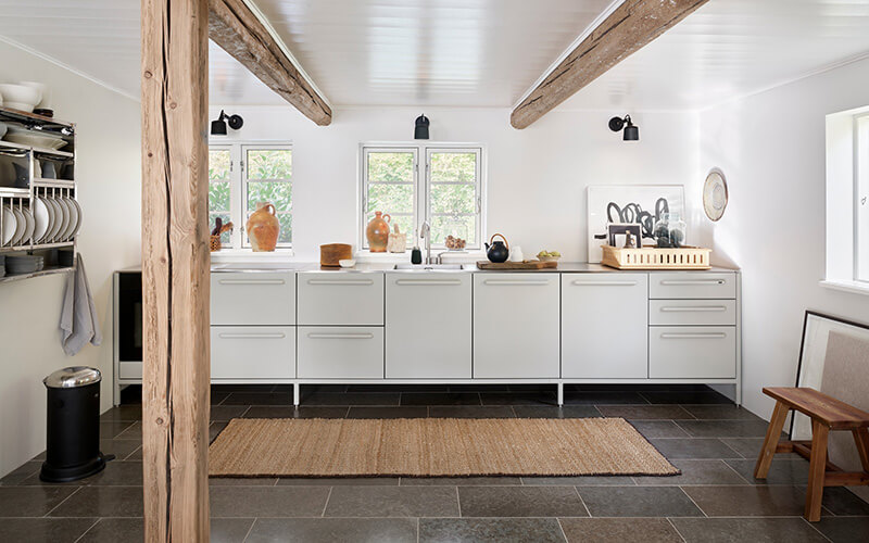 Helle Küche mit weißen Schränken, Holzdecke mit sichtbaren Balken, zwei Fenstern und einem Teppich auf dunklem Boden.