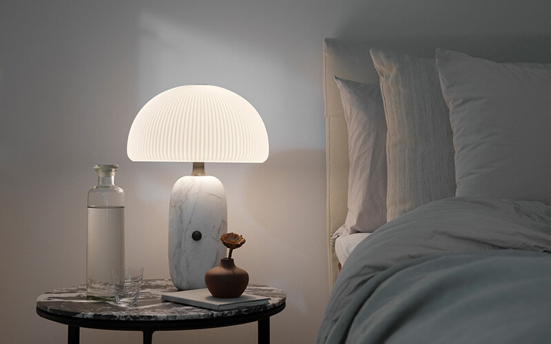 Marmorierter Nachttisch mit brauner Vase und beleuchteter Pilzlampe neben einem Bett mit Kissen und Decke.