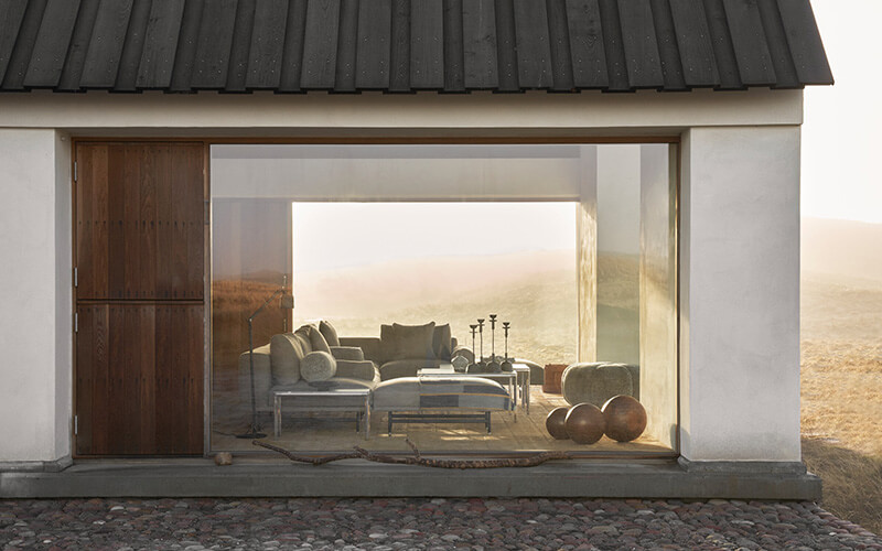 Modernes Ferienhaus mit großer Glasfront, das einen Blick auf eine minimalistisch aber gemütlich eingerichtete Sitzecke mit Sofa, Hockern und Kerzenhaltern bietet.