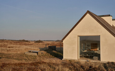 Modernes Ferienhaus mit großem Fenster, das in eine offene Landschaft mit trockenem Gras und leicht hügeligem Gelände blickt.