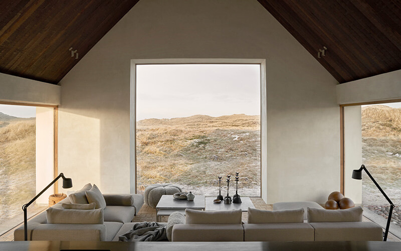 Wohnzimmer mit großen Fenstern, Sofa in U-Form, zwei Stehlampen und Blick auf die Dünen.