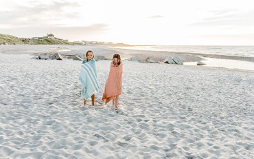 Zwei Kinder stehen auf einem Sandstrand, eingewickelt in gestreifte Handtücher, im Hintergrund Meer und Dünen.