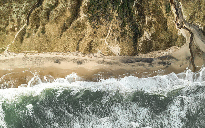 Luftaufnahme eines Strandes mit Wellen, Dünen und Pfaden im Sand hinter dem Strand.