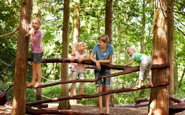 Vier Kinder klettern und lehnen an einem hölzernen Geländer zwischen Bäumen auf einem Waldspielplatz.