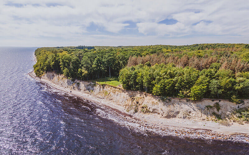 Klippenküste mit bewaldetem Plateau und felsigem Strand an einem sonnig-bewölkten Tag.