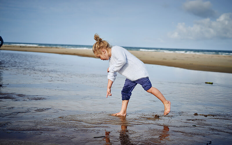 Kind in weißem Hemd und hochgekrempelter, blauer Hose zwigt auf etwas im nassen Sand am Strand.